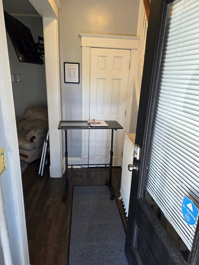 Narrow entryway with white six-panel door, black standing desk, dark hardwood floor, gray runner rug, and white blinds on window