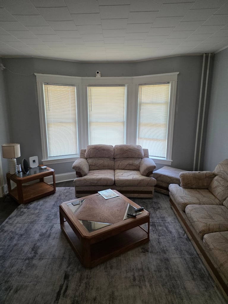 A contemporary living room with gray walls, beige furniture, bay windows with blinds, wooden coffee table, and a patterned area rug