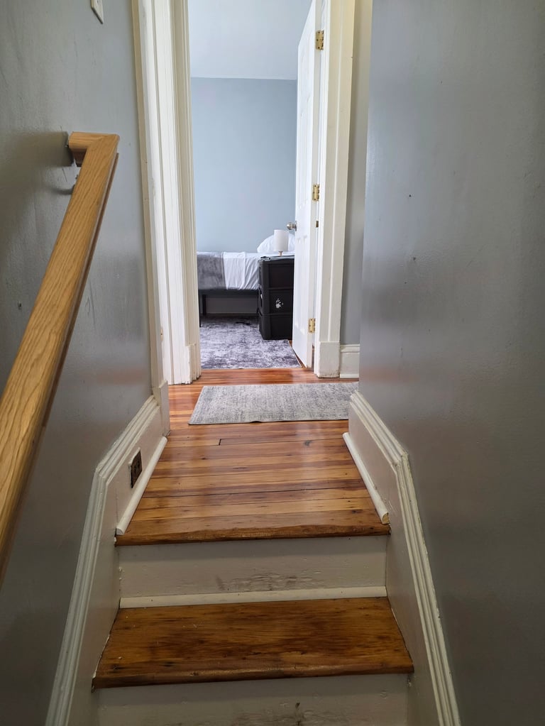 Interior staircase with wooden steps leading down to a hallway, with white walls and white trim, opening to a bedroom with a bed visible through the doorway