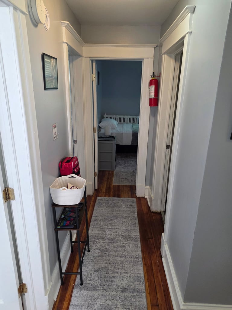 A narrow hallway with white walls and wood flooring, leading to a bedroom, with storage shelves and fire extinguisher on the left
