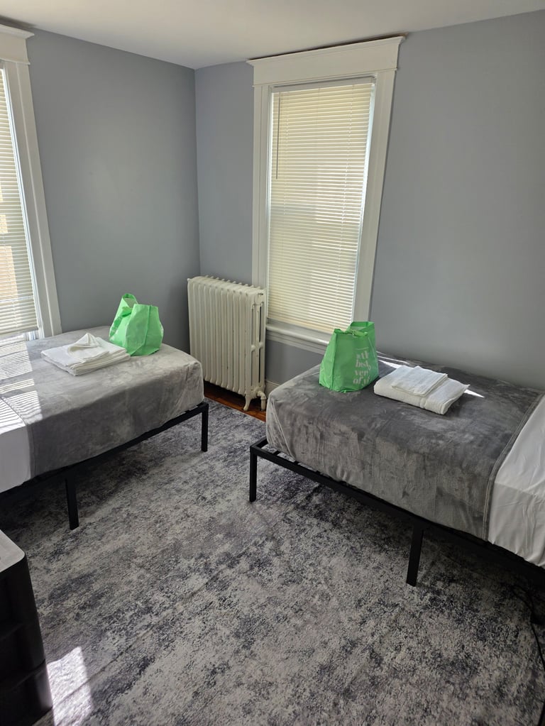 Sparse bedroom with two twin beds, white radiator, and windows with blinds