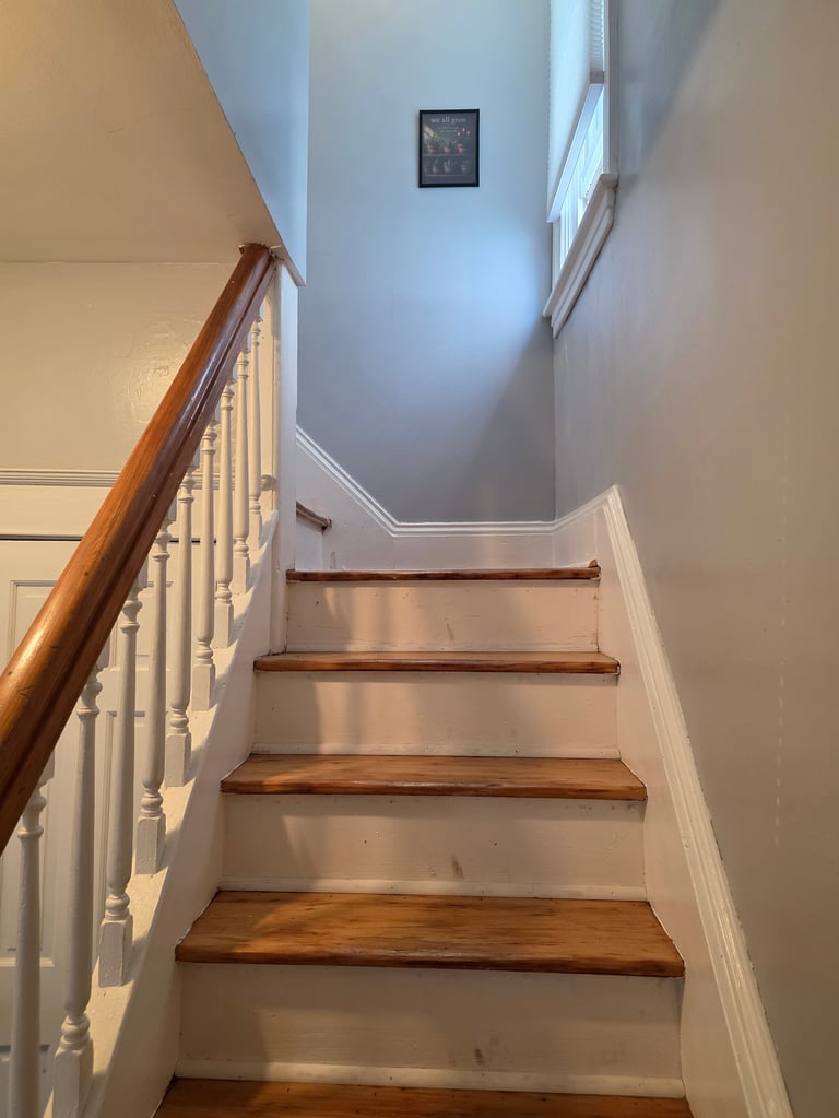 Interior staircase with wooden steps and brown handrail, white walls, and small clock on wall above