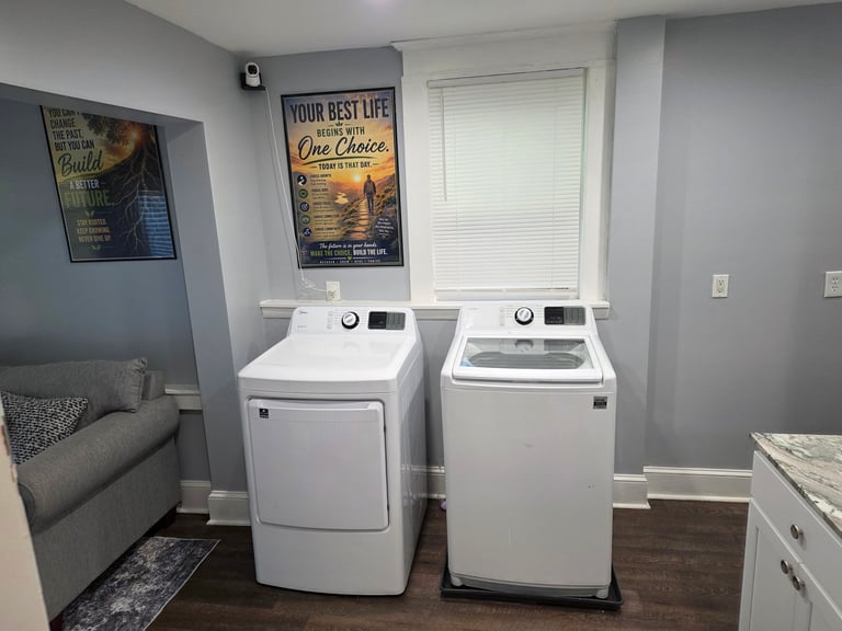 Laundry room with two white front-load washers side by side, inspirational wall signs, gray walls, and a couch in the corner