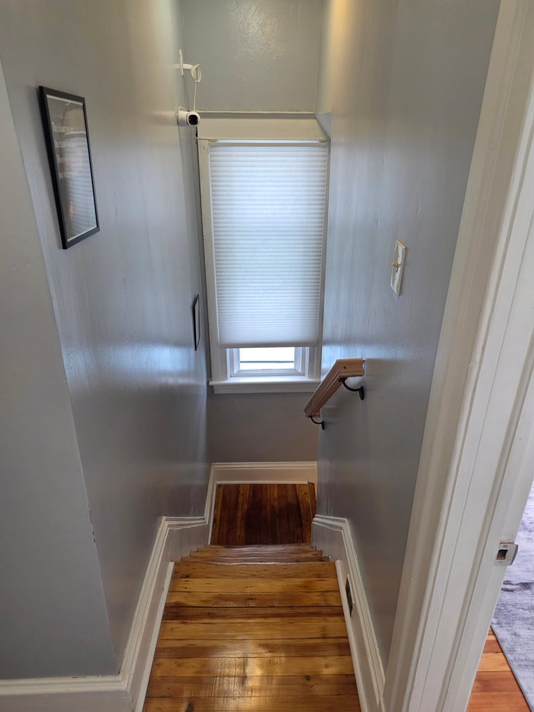 Narrow hallway with light gray walls, wooden flooring, and a window with blinds at the end