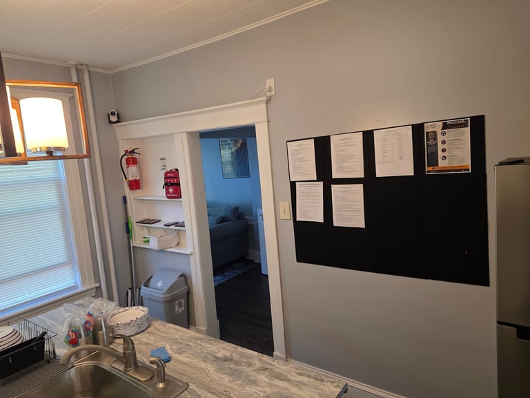 Office kitchen with white counter, window, doorway to blue-lit room, black bulletin board with papers, fire extinguishers, and refrigerator