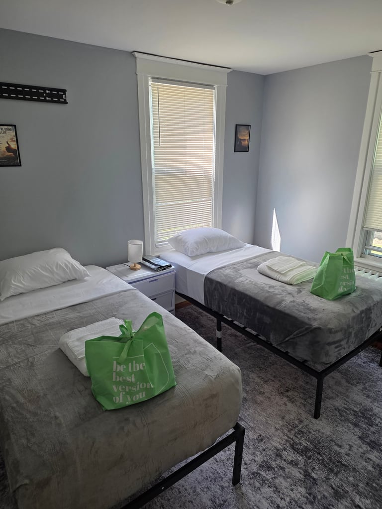 A clean dorm room with three single beds with white pillows and gray bedding, green welcome bags on the beds, gray walls, and bright windows with blinds