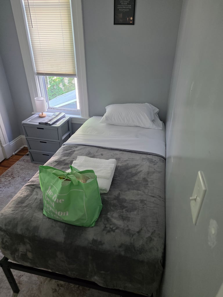 Minimalist bedroom with a single bed, gray blanket, white pillow, green reusable bag on bed, nightstand with items, window with blinds, and light green walls